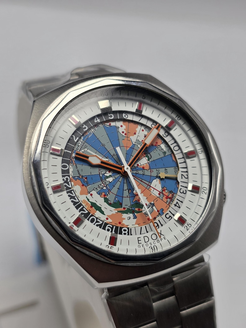 Op de afbeelding: Een roestvrijstalen EDOX Geoscope horloge met een wereldkaartontwerp op de wijzerplaat. Het horloge heeft een witte wijzerplaat met zwarte en rode markeringen, een datumweergave en een zilveren band. Het horloge heeft een unieke, vintage uitstraling.