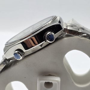 Op de afbeelding: Close-up van een zilveren polshorloge met een zwarte wijzerplaat en een zilveren band. Het horloge heeft twee blauwe knoppen aan de zijkant. Het horloge staat op een witte ondergrond.