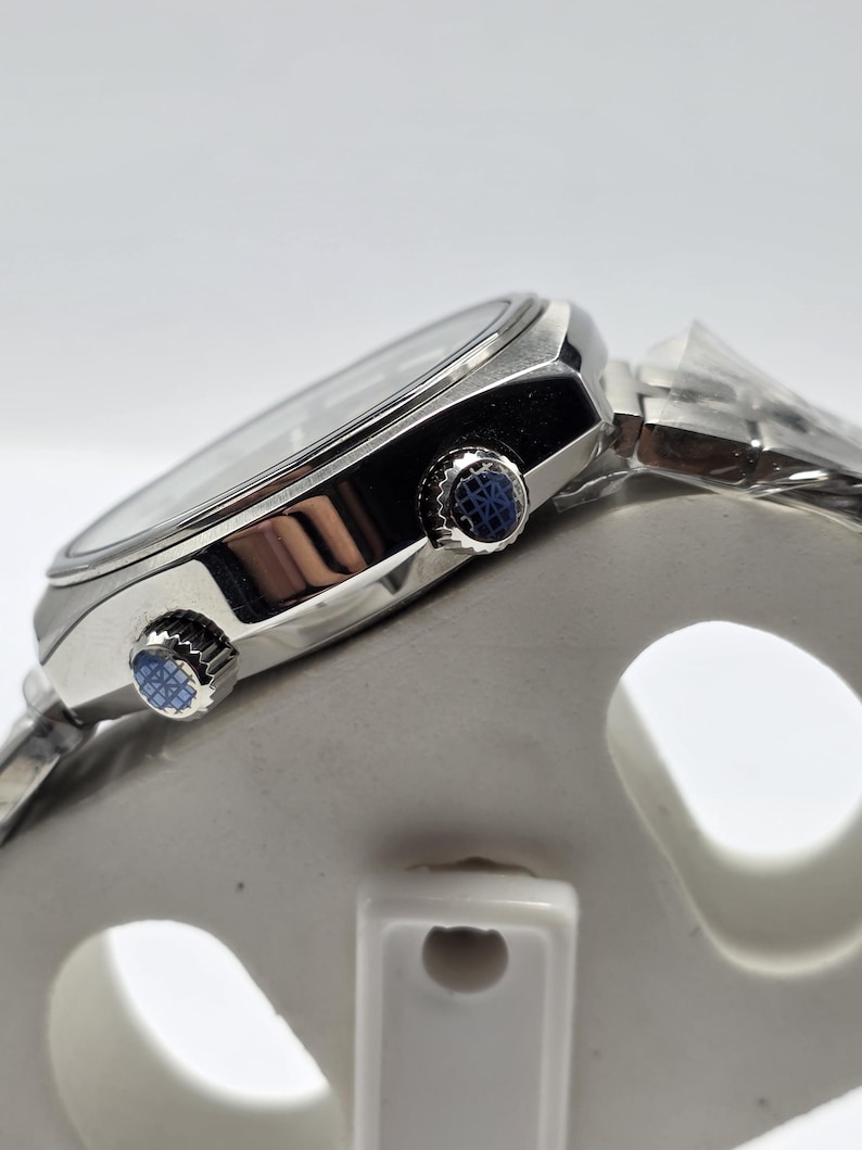 Op de afbeelding: Close-up van een zilveren polshorloge met een zwarte wijzerplaat en een zilveren band. Het horloge heeft twee blauwe knoppen aan de zijkant. Het horloge staat op een witte ondergrond.