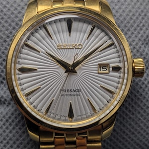 May include: Gold-toned Seiko Presage automatic wristwatch. The watch features a silver dial with a radial pattern, gold hands and markers, and a date window at the 3 o'clock position. The watch band is also gold-toned. The watch is made in Japan.