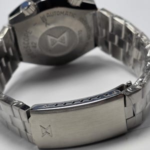 Op de afbeelding: Close-up van een roestvrijstalen polshorloge met een metalen band. Het horloge heeft een zilveren sluiting met een logo. De wijzerplaat is zichtbaar, met het woord "AUTOMATIC" en andere tekst. De band is gemaakt van kleine, rechthoekige metalen schakels.