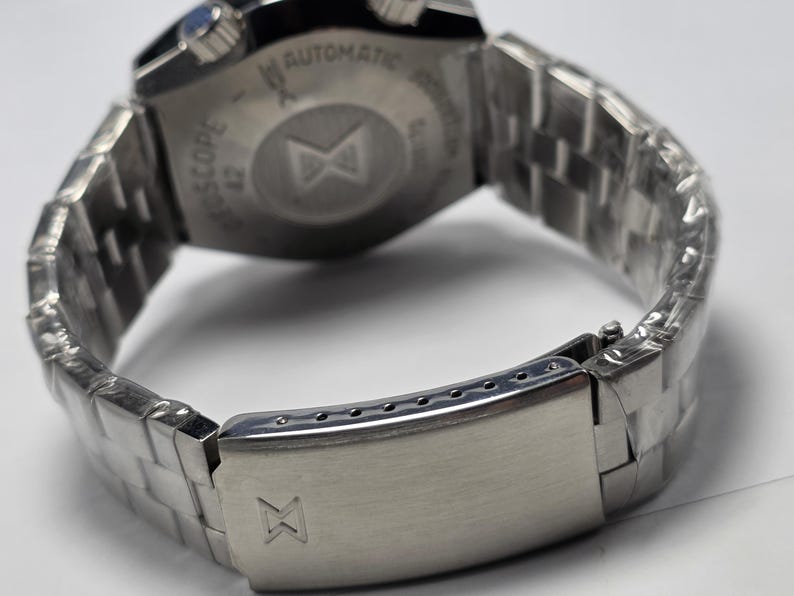 Op de afbeelding: Close-up van een roestvrijstalen polshorloge met een metalen band. Het horloge heeft een zilveren sluiting met een logo. De wijzerplaat is zichtbaar, met het woord "AUTOMATIC" en andere tekst. De band is gemaakt van kleine, rechthoekige metalen schakels.