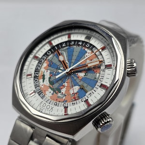 Op de afbeelding: Een zilverkleurig horloge met een witte wijzerplaat met een wereldkaart in blauw, oranje en groen. Het horloge heeft een roestvrijstalen band en een getextureerde lunette. De wijzerplaat bevat de tekst "EDOX GEOSCOPE 30".