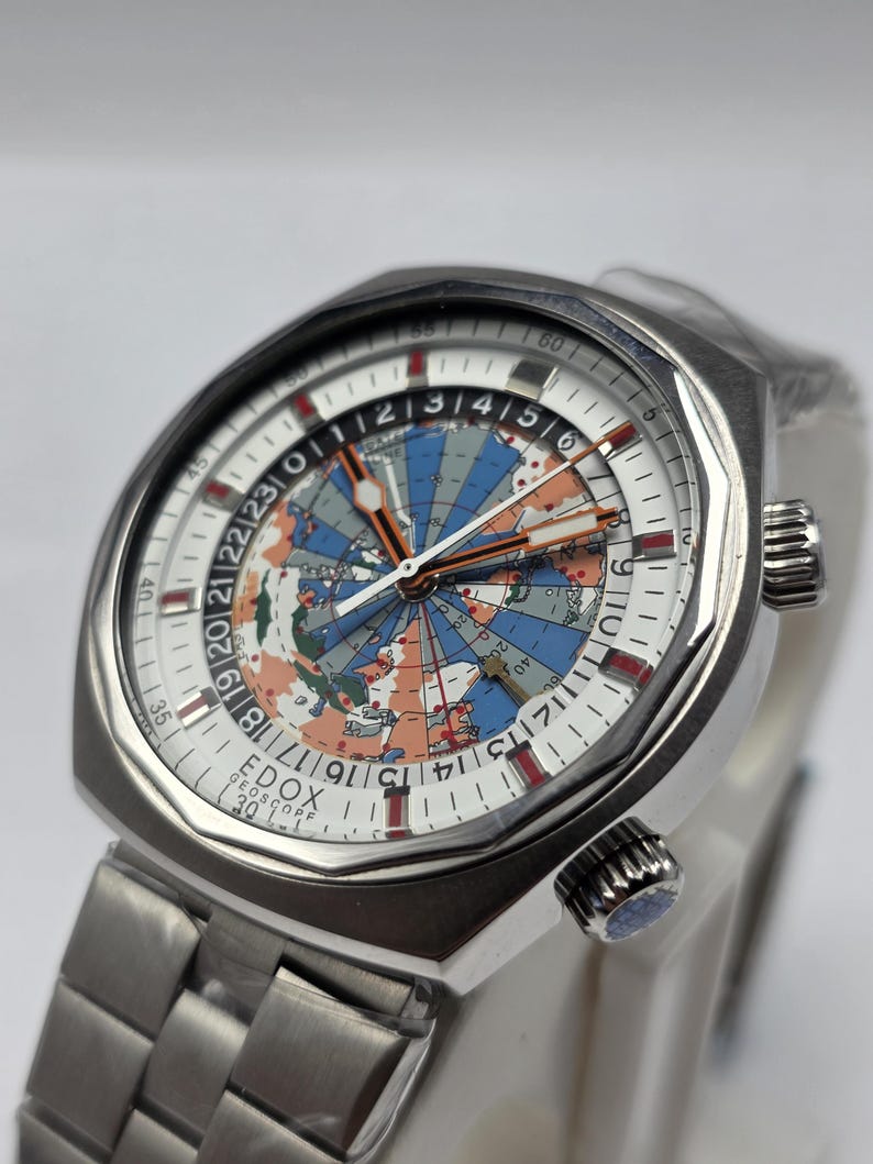 Op de afbeelding: Een zilverkleurig horloge met een witte wijzerplaat met een wereldkaart in blauw, oranje en groen. Het horloge heeft een roestvrijstalen band en een getextureerde lunette. De wijzerplaat bevat de tekst "EDOX GEOSCOPE 30".