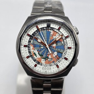 Op de afbeelding: Een roestvrijstalen polshorloge met een witte wijzerplaat met een wereldkaartontwerp. Het horloge heeft een zilveren band en een zwarte lunette. De wijzerplaat toont de merknaam "EDOX GEOSCOPE".