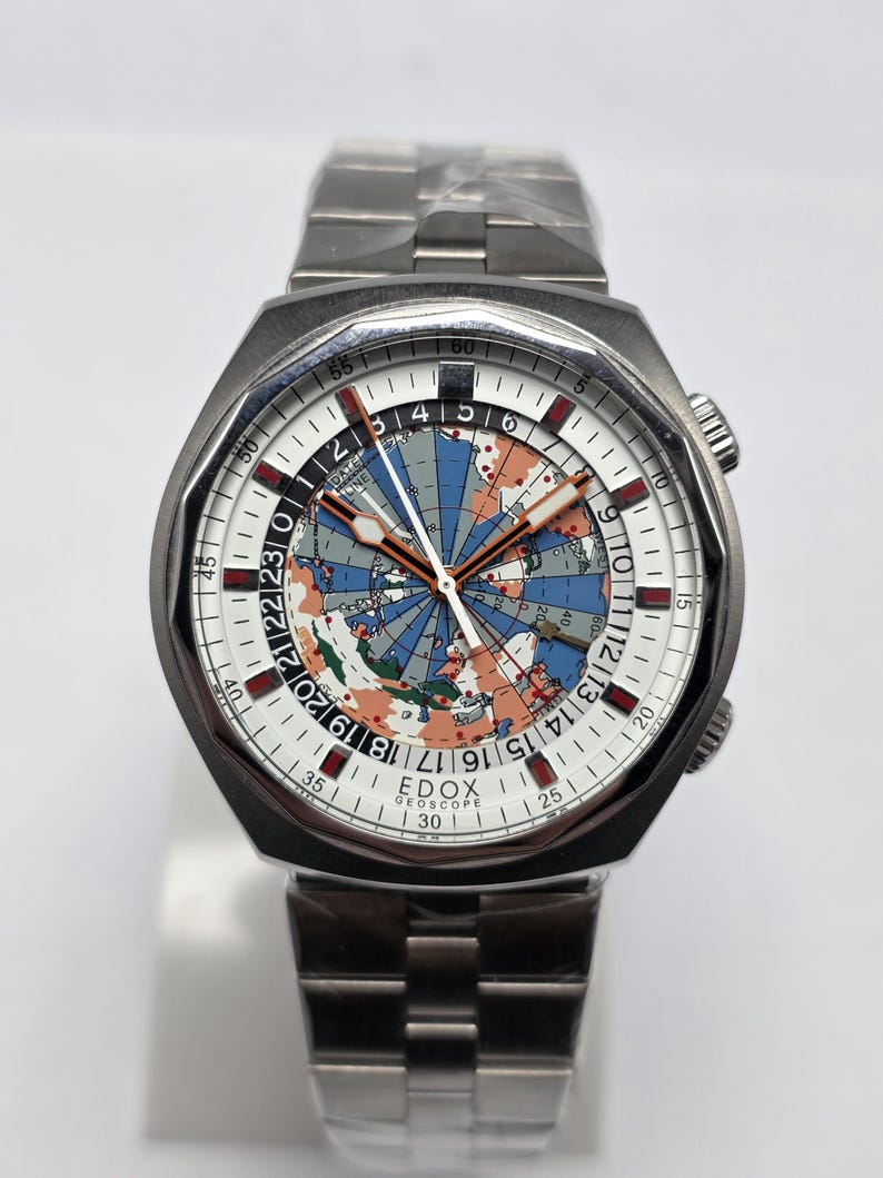 Op de afbeelding: Een roestvrijstalen polshorloge met een witte wijzerplaat met een wereldkaartontwerp. Het horloge heeft een zilveren band en een zwarte lunette. De wijzerplaat toont de merknaam "EDOX GEOSCOPE".