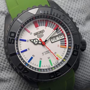May include: A black Seiko 5 automatic watch with a white face, colorful markers, and a green rubber strap. The watch features a black bezel with white numerals and the day of the week displayed at the 3 o'clock position.