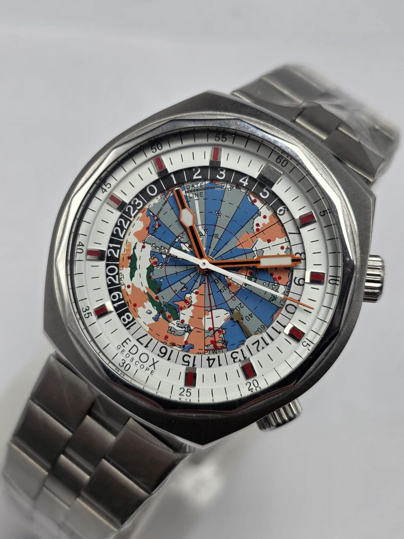 Op de afbeelding: Een roestvrijstalen polshorloge met een wereldkaartontwerp op de wijzerplaat. Het horloge heeft een witte wijzerplaat met zwarte en rode markeringen en een zilveren band. De wijzerplaat toont de tekst "EDOX GEOSCOPE".