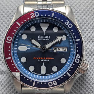 May include: A stainless steel Seiko automatic diver's watch with a blue and red rotating bezel. The watch face is blue with white markers and hands, and the date is displayed at the 3 o'clock position. The watch is marked "Diver's 200m" and "Made in Japan."