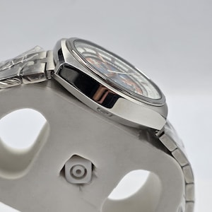 Op de afbeelding: Een zilverkleurig polshorloge met een patroon op de wijzerplaat in blauwe en oranje tinten. Het horloge heeft een roestvrijstalen band en wordt gepresenteerd op een witte standaard. De wijzerplaat wordt beschermd door een helder kristal.