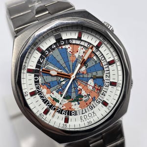 Op de afbeelding: Zilverkleurig polshorloge met een witte wijzerplaat met een wereldkaartontwerp in blauw, oranje en grijs. Het horloge heeft een roestvrijstalen band. De tekst "EDOX GEOSCOPE" is zichtbaar op de wijzerplaat.