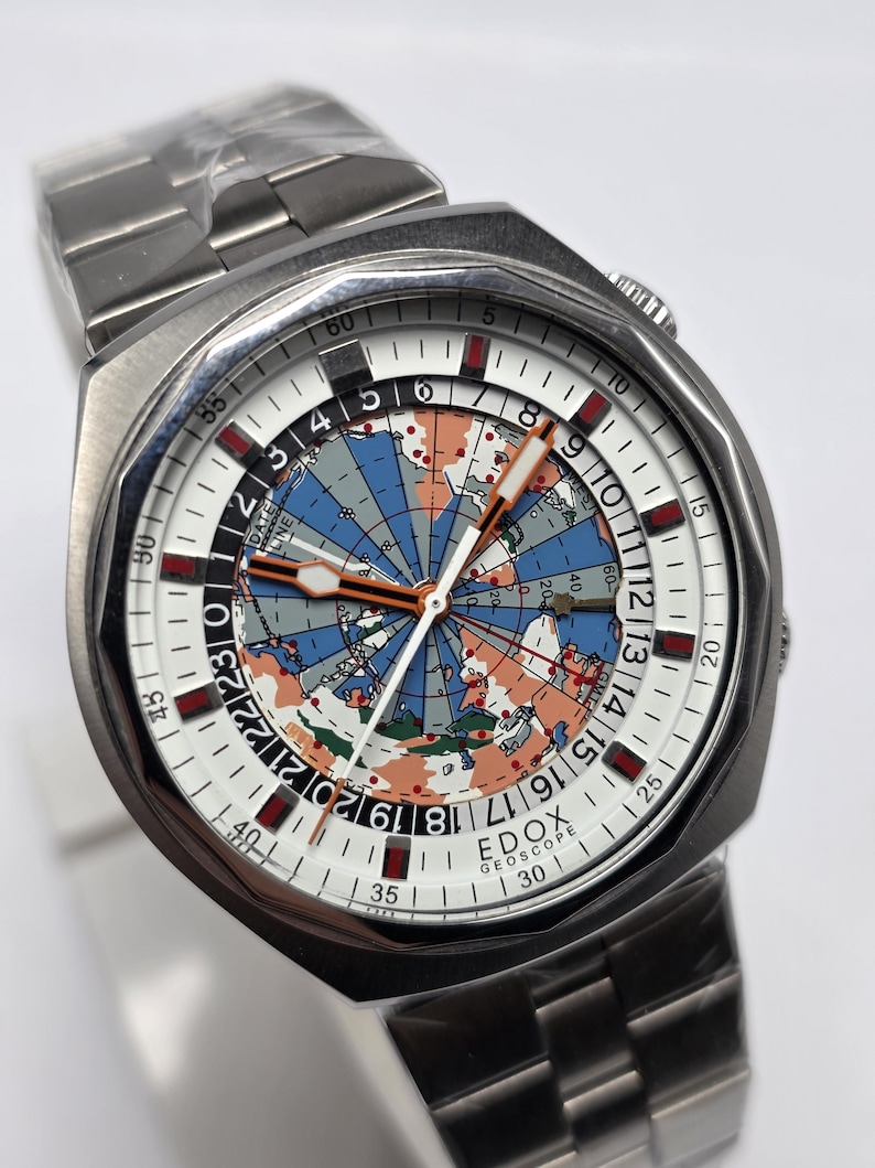Op de afbeelding: Zilverkleurig polshorloge met een witte wijzerplaat met een wereldkaartontwerp in blauw, oranje en grijs. Het horloge heeft een roestvrijstalen band. De tekst "EDOX GEOSCOPE" is zichtbaar op de wijzerplaat.