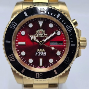May include: A gold-tone stainless steel wristwatch featuring a deep red sunburst dial, a black rotating bezel with white markers, and luminous hour indices. The dial displays the Orient logo, AAA Crystal, 21 Jewels, and a day-date window.
