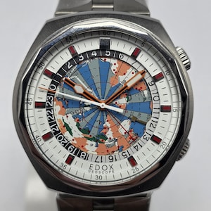 May include: A stainless steel Edox Geoscope watch with a white dial featuring a world map design. The watch has a silver band and a rotating bezel. The dial includes orange and red accents, with a date line indicator.