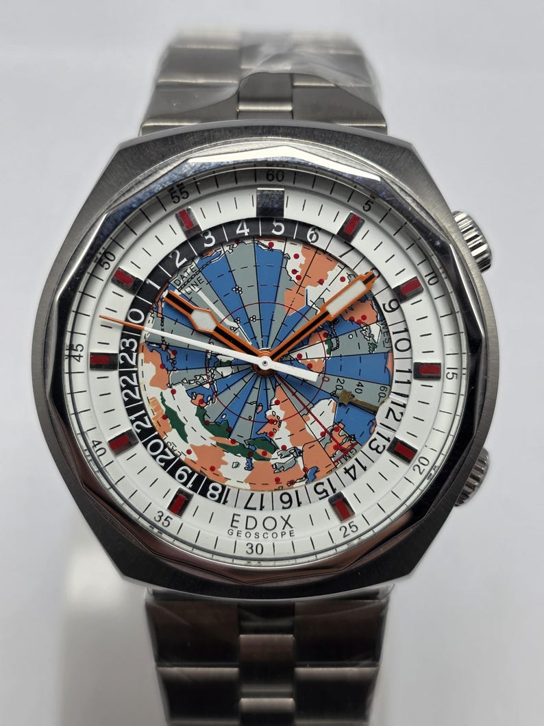 Op de afbeelding: Een roestvrijstalen Edox Geoscope horloge met een witte wijzerplaat met een wereldkaartontwerp. Het horloge heeft een zilveren band en een draaibare lunette. De wijzerplaat bevat oranje en rode accenten, met een datumlijn.