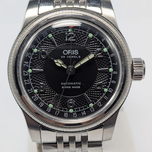 May include: A silver Oris wristwatch with a black dial and a stainless steel bracelet. The watch face has luminous hands and markers, a date display, and the text "ORIS 25 JEWELS AUTOMATIC SWISS MADE". The watch has a fluted bezel and a crown.