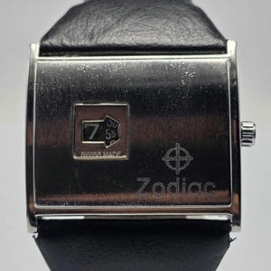 May include: A silver-toned Zodiac watch with a black leather band. The watch face displays the time as 7:55 and the words "Swiss Made." The Zodiac logo is visible below the time display.
