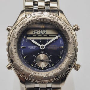 May include: Silver-toned Casio Mission watch with a blue face. Features include a digital display showing 12:00, multiple dials, and text indicating chronograph functions. The watchband is a matching silver link style.