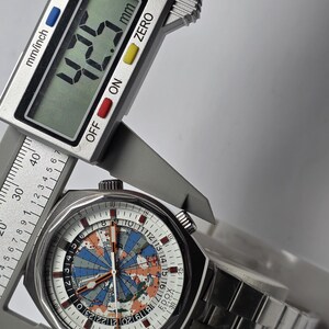 Op de afbeelding: Een roestvrijstalen polshorloge met een witte wijzerplaat met een wereldkaartontwerp in blauw, oranje en grijs. Het horloge wordt gemeten met een digitale schuifmaat, met een breedte van 42,5 mm.
