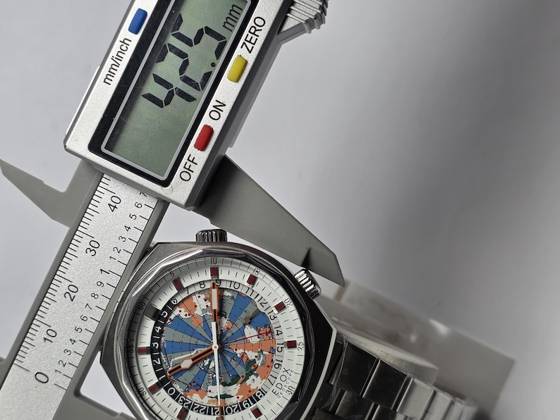 Op de afbeelding: Een roestvrijstalen polshorloge met een witte wijzerplaat met een wereldkaartontwerp in blauw, oranje en grijs. Het horloge wordt gemeten met een digitale schuifmaat, met een breedte van 42,5 mm.