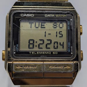 Puede incluir: Un reloj digital Casio Data Bank Telememo 50 en tono dorado. La esfera del reloj muestra la fecha y la hora: martes 30, 1-15 y 8:22:04. El reloj tiene una correa de metal dorada y botones con las etiquetas "REVERSE" y "FORWARD".