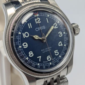 Może przedstawiać: Zegarek automatyczny Oris Big Crown ze stali nierdzewnej z ciemnoniebieską tarczą. Zegarek posiada białe cyfry arabskie, świecące wskazówki i wskaźnik daty. Pasek to bransoleta ze stali nierdzewnej.