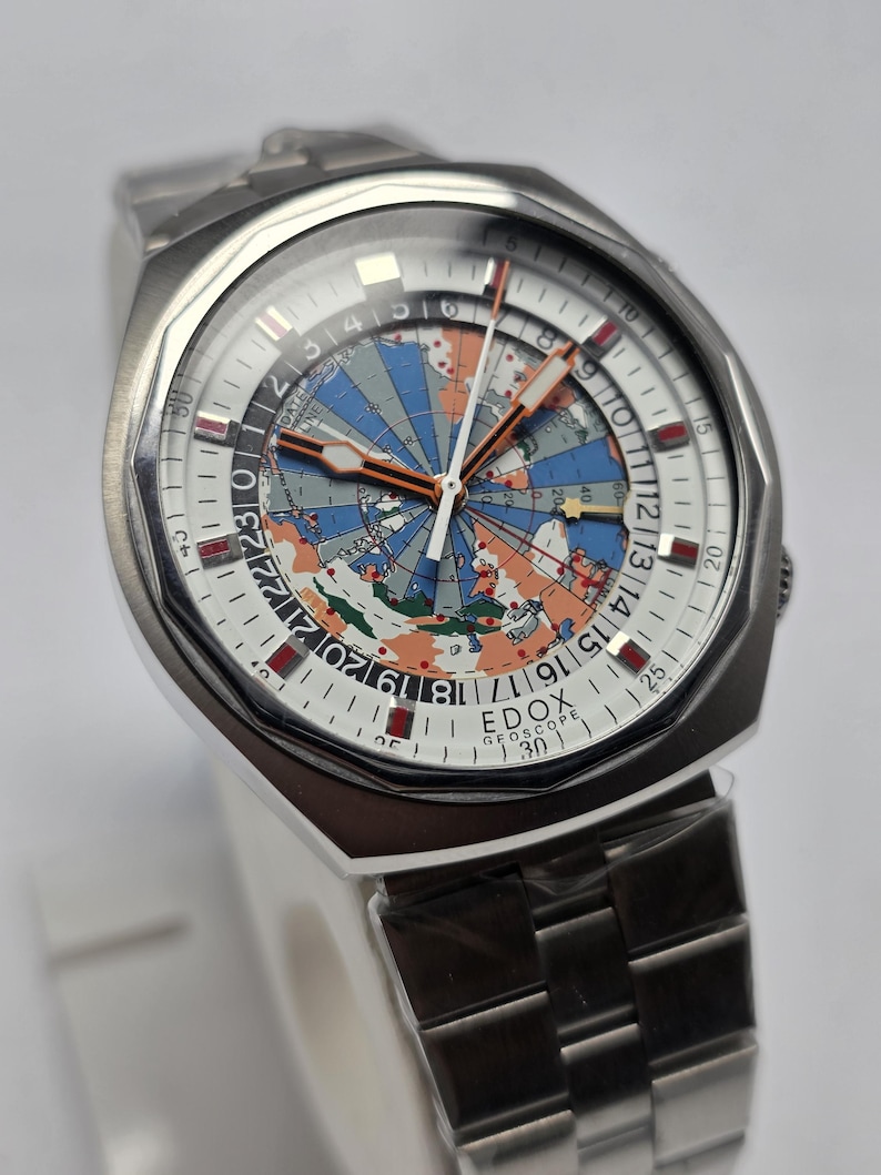 Op de afbeelding: Een roestvrijstalen Edox Geoscope horloge met een witte wijzerplaat met een wereldkaartontwerp in blauw, oranje en groen. Het horloge heeft een zilveren band en een datumweergave.