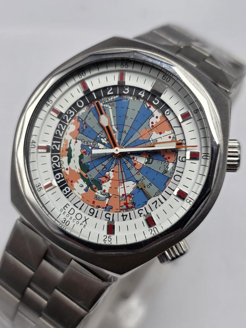 Op de afbeelding: Een roestvrijstalen polshorloge met een witte wijzerplaat met een wereldkaartontwerp in blauw, oranje en groen. Het horloge heeft een zilveren band en de merknaam "EDOX GEOSCOPE" is zichtbaar. De diameter van de wijzerplaat is ongeveer 4 cm.