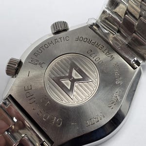 Op de afbeelding: Een Geoscope automatisch polshorloge van roestvrij staal. De achterkant van het horloge heeft de tekst "WATERPROOF", "SUPER SWISS" en "MADE". Het horloge heeft een zilveren band en een getextureerde kroon. Het horloge zit nog in plastic verpakt.