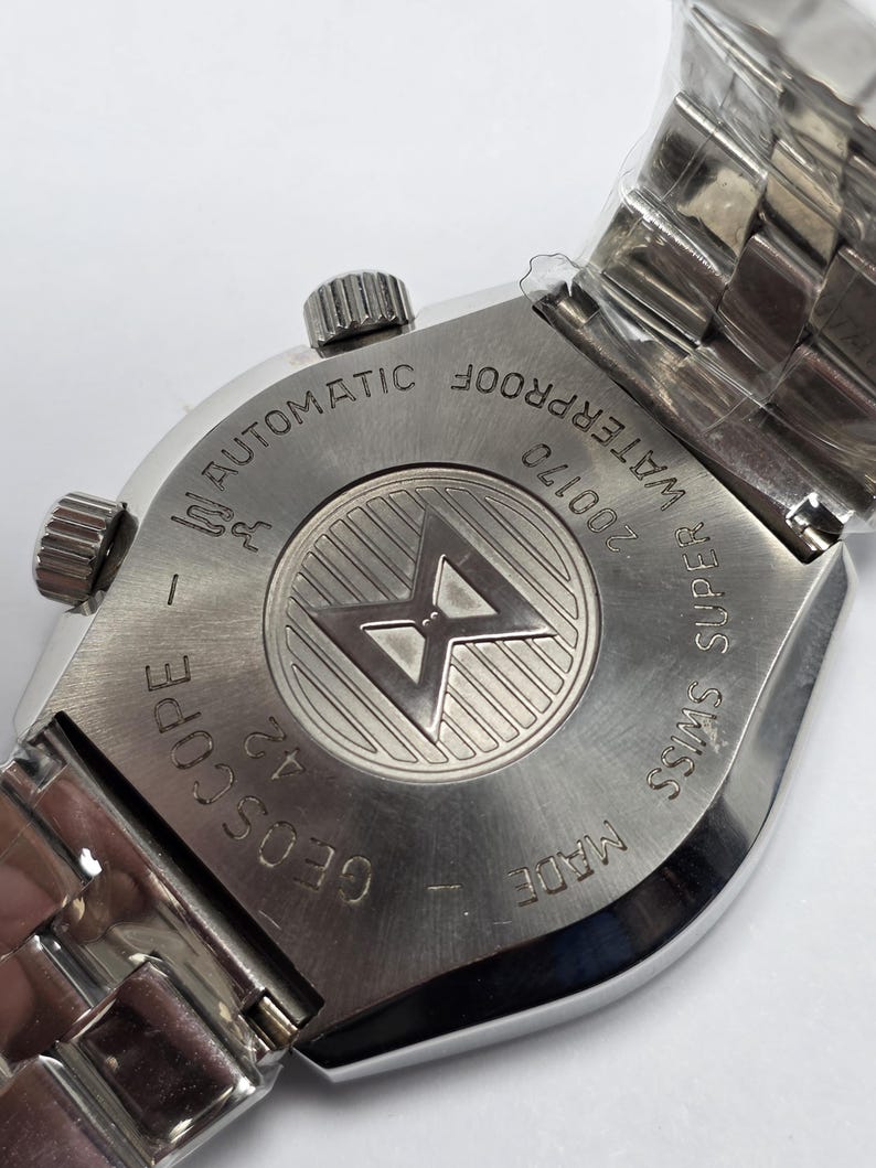 Op de afbeelding: Een Geoscope automatisch polshorloge van roestvrij staal. De achterkant van het horloge heeft de tekst "WATERPROOF", "SUPER SWISS" en "MADE". Het horloge heeft een zilveren band en een getextureerde kroon. Het horloge zit nog in plastic verpakt.