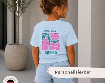 May include: A light blue short-sleeved t-shirt featuring a graphic on the back with pink flowers, teal leaves, and the text COOL WILD SIEBEN. The shirt is paired with light blue denim shorts. The design includes a central floral motif.