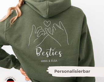 May include: An olive green hooded sweatshirt featuring a white line art graphic of two hands making a pinky promise with a heart above them. Below the graphic, the word Besties is printed in a script font, followed by the text ANNA & ELSA.