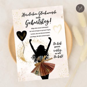 May include: A birthday greeting card with German text and a silhouette of a person with arms raised. The person's skirt is made of folded Euro banknotes. A black heart balloon and gold accents decorate the card.