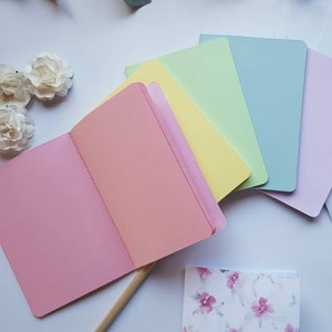 May include: A set of six pastel coloured notebooks, including pink, peach, yellow, light green, light blue, and lavender. The notebooks are arranged in a rainbow pattern and are open to reveal blank pages.
