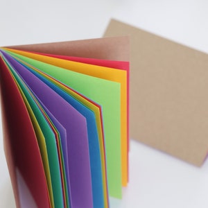 May include: A stack of rainbow-coloured paper, folded in half, with a brown paper cover. The paper is arranged in a rainbow pattern, with red, orange, yellow, green, blue, indigo, and violet.