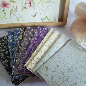 May include: A set of ten small, decorative paper notebooks with a floral pattern. The notebooks are arranged in a fan shape, with the lightest colour at the top and the darkest colour at the bottom. The notebooks are all the same size and have a silver floral pattern.