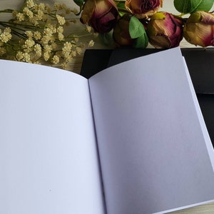 May include: A black notebook with a white interior, open to a blank page. The notebook is lying on a wooden surface with a bouquet of pink and white flowers in the background.