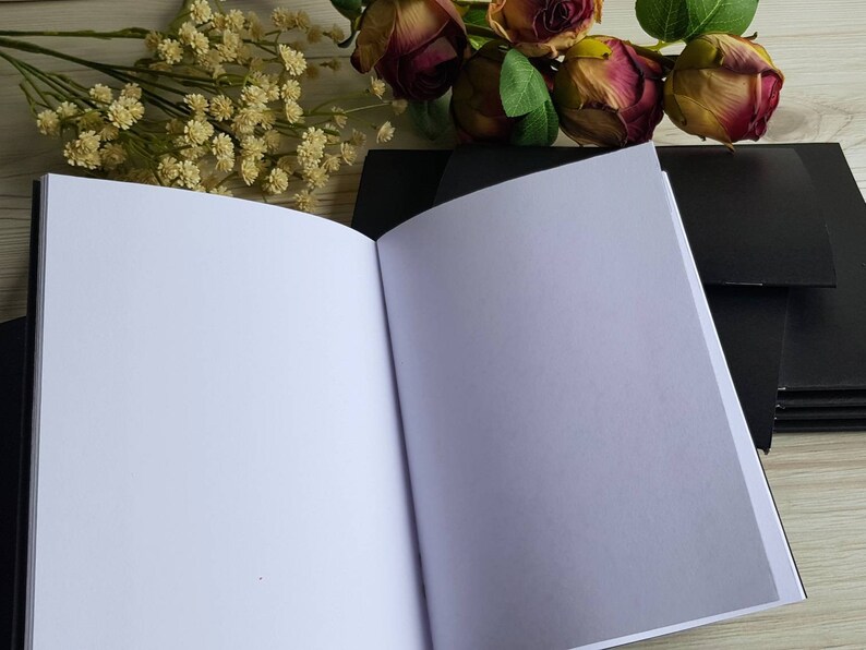 May include: A black notebook with a white interior, open to a blank page. The notebook is lying on a wooden surface with a bouquet of pink and white flowers in the background.