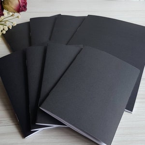 May include: A stack of eight black notebooks with white edges. The notebooks are arranged in a fan shape, with the top notebook slightly overlapping the others.