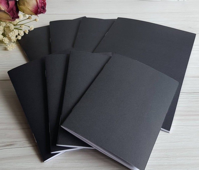 May include: A stack of eight black notebooks with white edges. The notebooks are arranged in a fan shape, with the top notebook slightly overlapping the others.