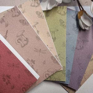 May include: A set of rectangular paper bookmarks in various colours including red, tan, olive green, blue, and purple. Each bookmark features a repeating line art pattern of mushrooms, dragonflies, and leaves printed in a dark brown ink.