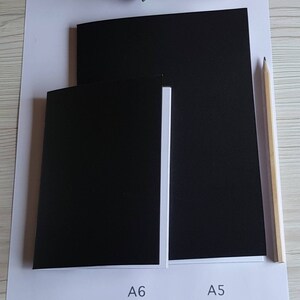 May include: Two black paper notebooks, one labeled "A6" and the other labeled "A5", are shown side-by-side on a white surface. A wooden pencil is lying on the surface to the right of the notebooks.
