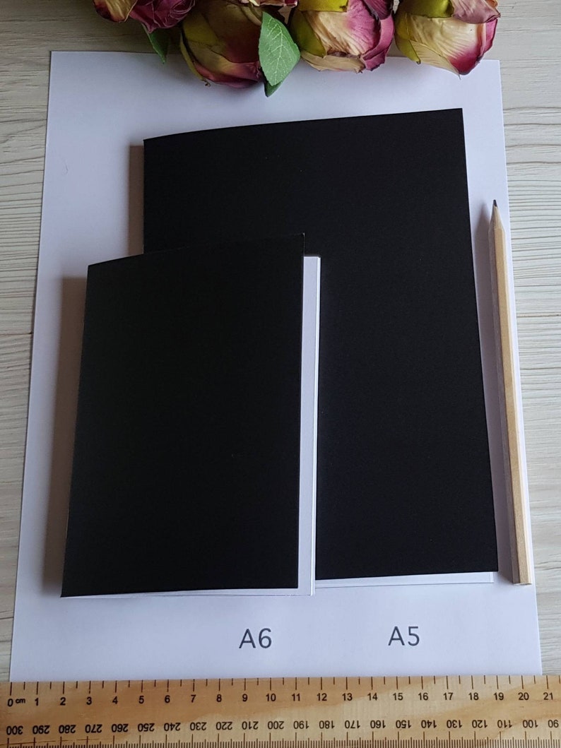 May include: Two black paper notebooks, one labeled "A6" and the other labeled "A5", are shown side-by-side on a white surface. A wooden pencil is lying on the surface to the right of the notebooks.