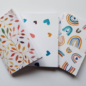 May include: Three notebooks with watercolour designs. One features colourful leaves, another has blue and orange hearts, and the third displays rainbows. The notebooks are white with the designs on the covers.