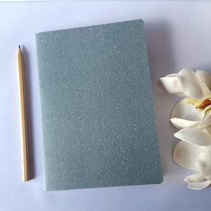 May include: A grey notebook and a wooden pencil rest on a white surface, with a white flower in the background. The notebook has a textured cover. The pencil is sharpened and ready for use.