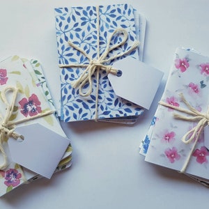 May include: Three stacks of small notebooks with floral designs. Each stack is tied with twine and has a blank tag attached. The designs include blue and white leaves, pink and white flowers, and red and white flowers.