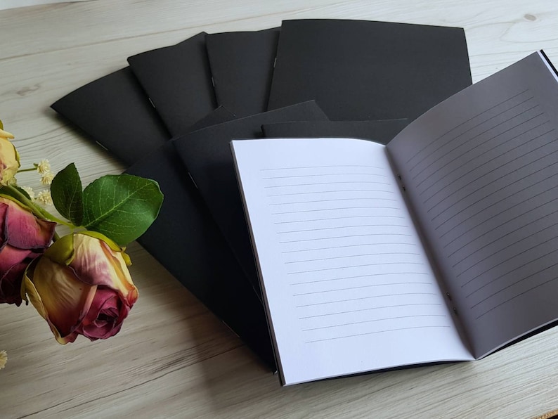 May include: A stack of black notebooks with lined pages. The top notebook is open, revealing the white lined paper inside.