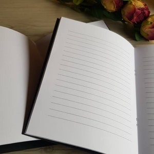 May include: Two black-bound notebooks with lined white pages. The notebooks are open and lying flat on a wooden surface.