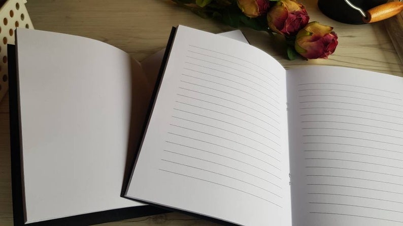 May include: Two black-bound notebooks with lined white pages. The notebooks are open and lying flat on a wooden surface.