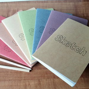 May include: A selection of colourful sketchbooks, including pink, beige, green, blue, purple, and brown. The word "Sketch" is printed on the front of the brown sketchbook. Each book has a simple "S" design on the cover.